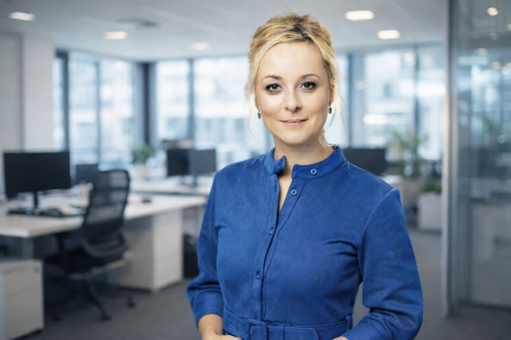 Frau in blauer Bluse steht in modernem Büro mit Computerarbeitsplätzen im Hintergrund.