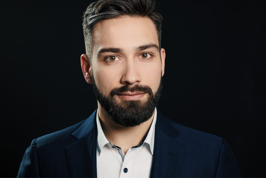 Mann mit kurzem Bart, Anzug und neutralem Gesichtsausdruck vor dunklem Hintergrund.