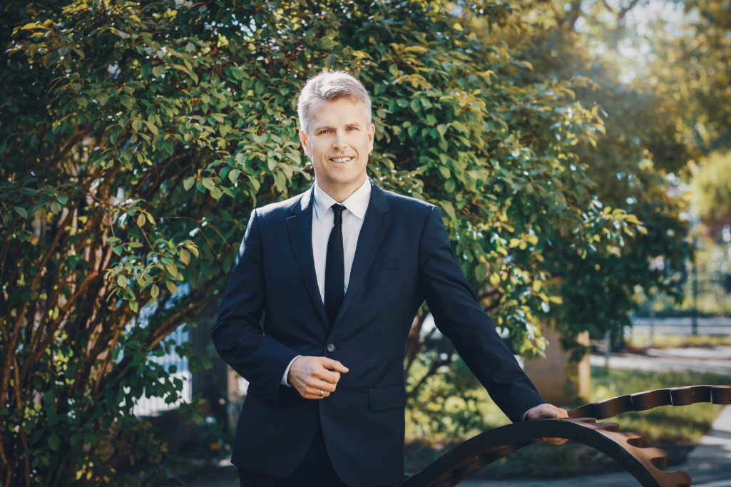 Mann in Anzug steht im Freien vor grünen Bäumen und lächelt.