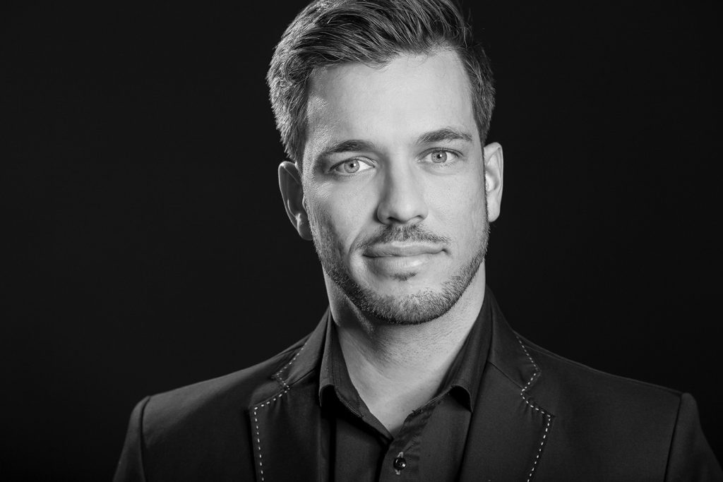 Porträt eines Mannes mit kurzem Haar, in einem schwarzen Anzug auf dunklem Hintergrund.