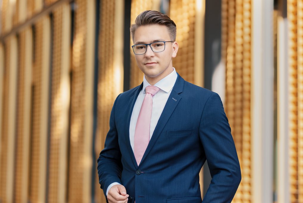 Junger Mann in Anzug mit Brille und Krawatte vor modernem Gebäudefond.