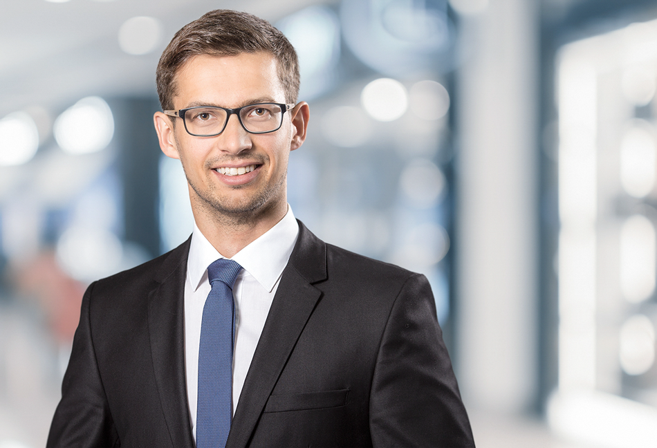 Mann in Anzug mit Brille, lächelt vor unscharfem urbanem Hintergrund.